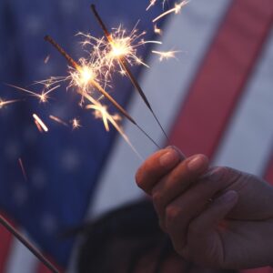 Two people holding sparklers in front of a American flag on the Fourth of July.