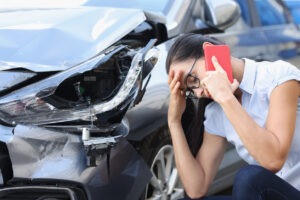 A woman on the phone squatting next to her totaled car from a car accident.