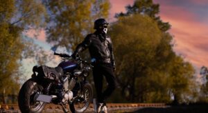 A motorcyclist in a leather jacket standing next to his motorcycle at sunset.