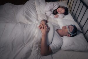 An asian man hooked up to a CPAP machine sleeping adjacent to his wife.