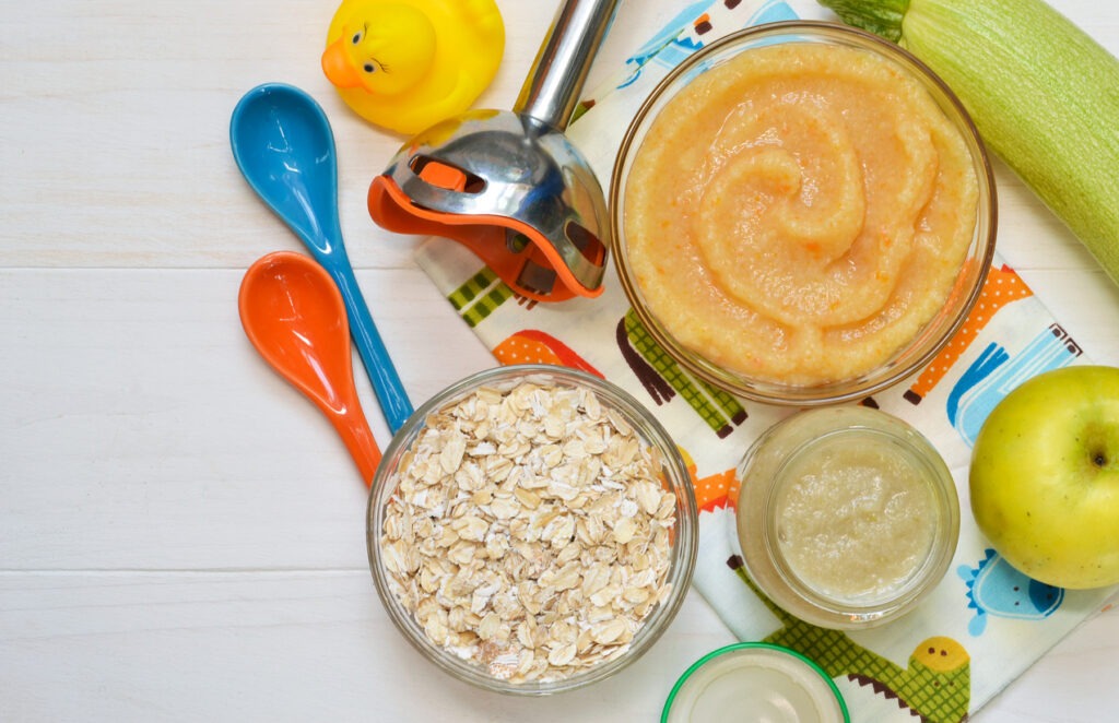 A table with different types of baby food in glass containers next to a container of oats and a green apple.