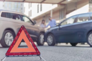 An orange triangular road safety sign in front of a car accident between two sedans.