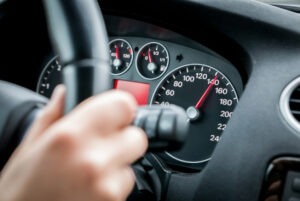 A closeup of a hand resting on a steering wheel in a car with the speedometer indicating a car speeding far above the speed limit.