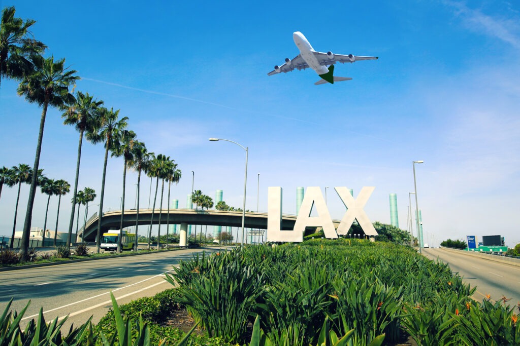 A plane flying overhead in a blue sky above a sign that says "LAX" outside of the LAX airport.