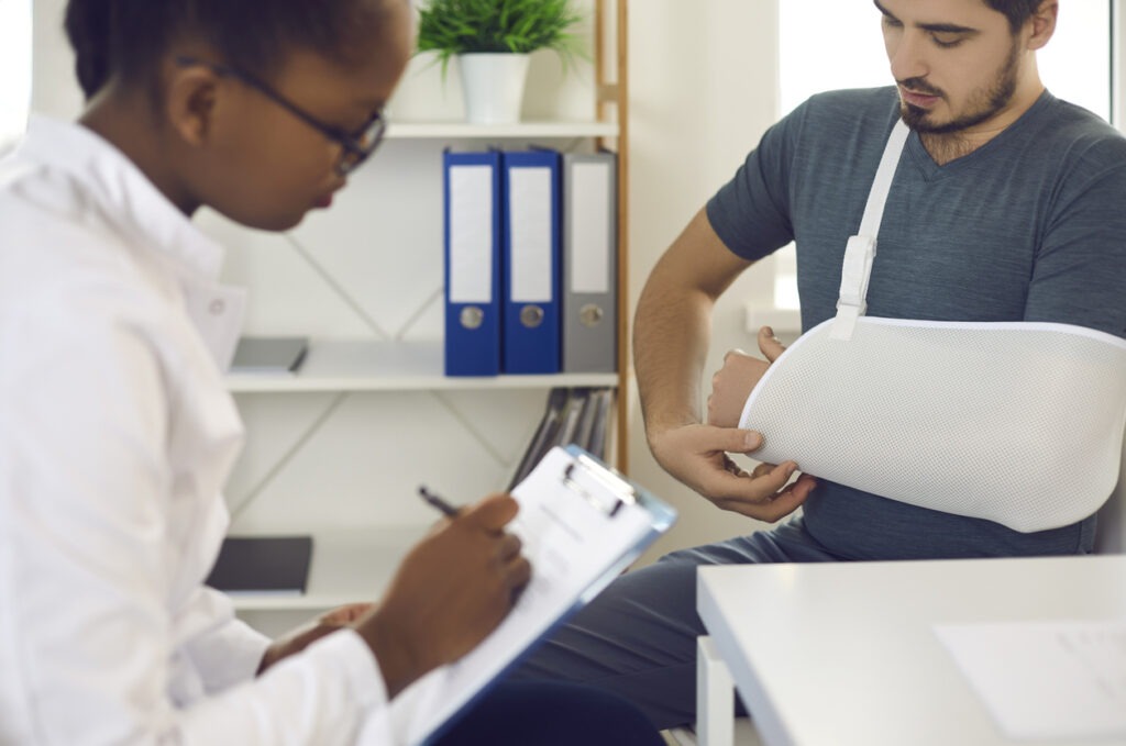 Young patient wearing injury support, forearm sling immobilizer, brace, cast, or scarf bandage on broken hand, wrist, elbow or arm seeing doctor at hospital.