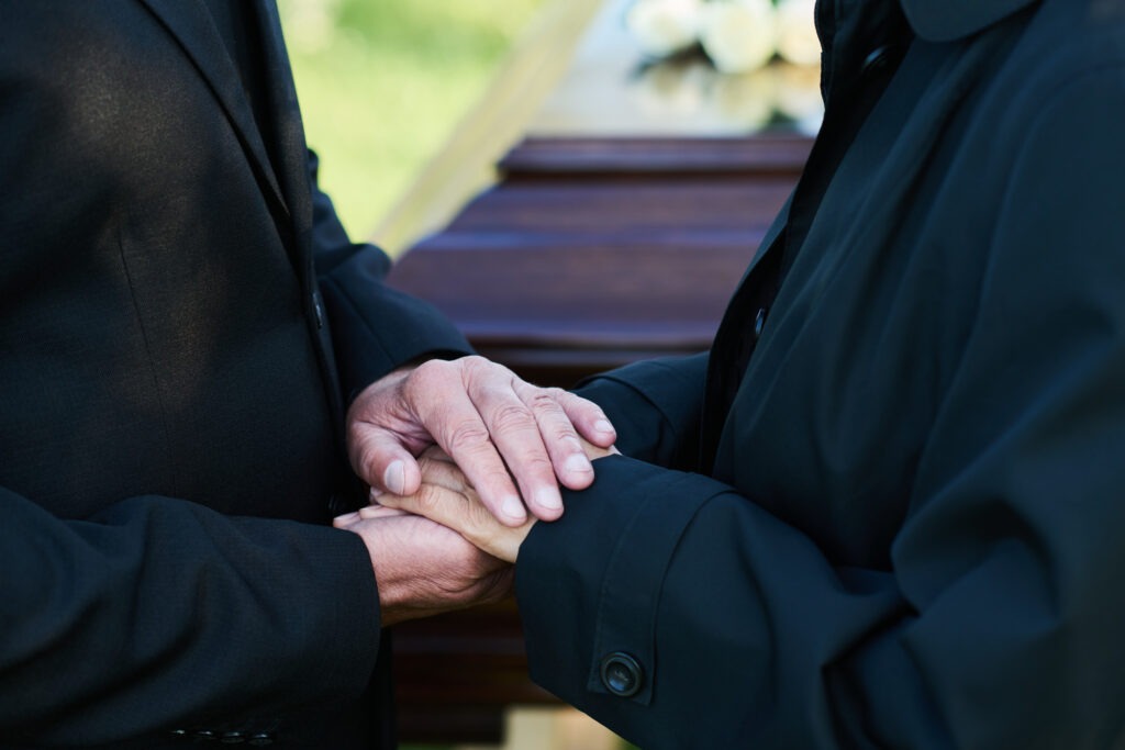 Two women holding hands in support with a casket behind them at a funeral on a sunny day.