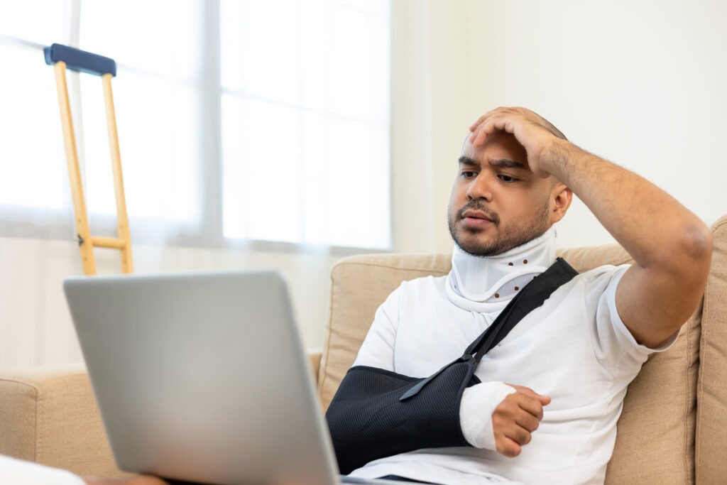 A man with an injured arm in a sling looking at his laptop in confusion.