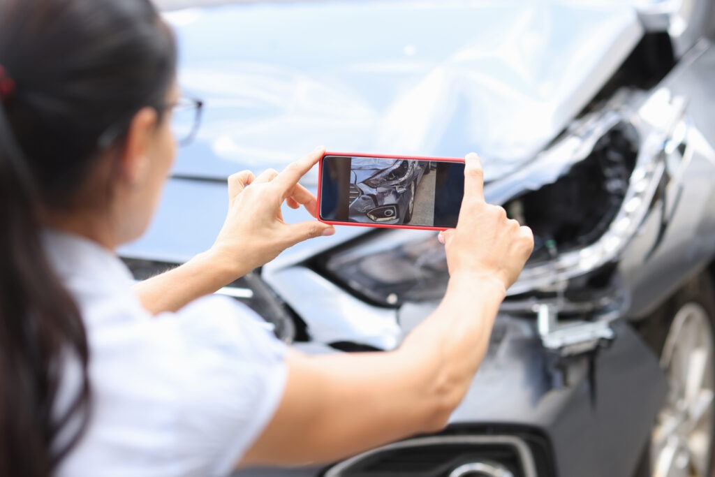 A young woman with a ponytail holding her red phone steady to take a photo of her damaged car.