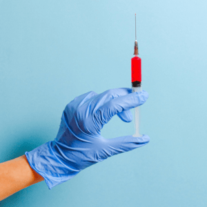 A blue-gloved hand holding a syringe in front of a baby blue backdrop.