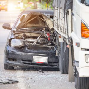 A totaled black car on the side of the road behind a white 18-wheeler truck.