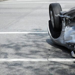 A silver car just involved in a car crash at an intersection of a city street, upside down and against the curb.