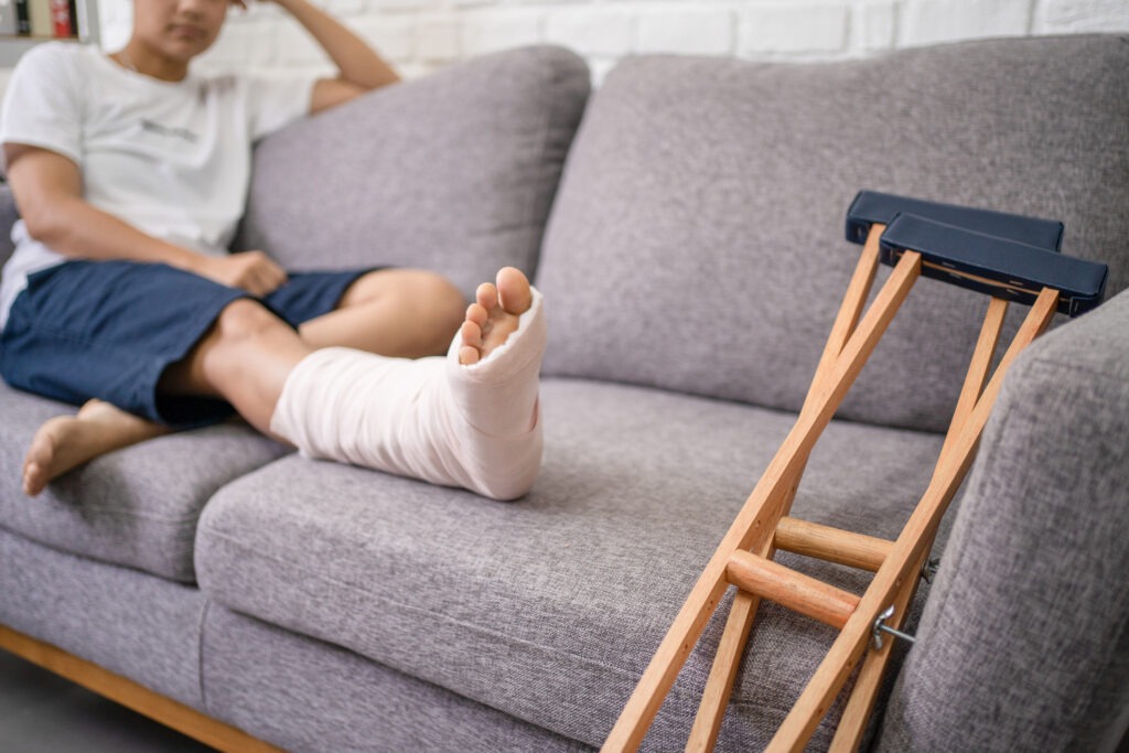 A young man with their leg in a cast resting on the couch next to their crutches.