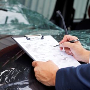 A man writing on a clipboard at the scene of a destructive car accident with a totaled car in the background.