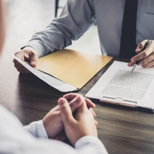 A lawyer in a suit with a folder in one hand and a clipboard in his other speaking to a client across from his wooden desk.