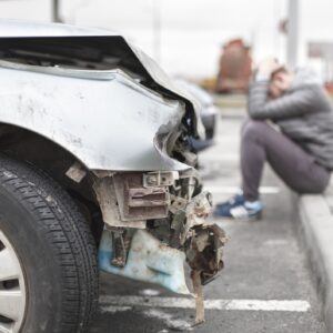 A totaled car on the side of the road with a man scrunched over stressed on the sidewalk nearby.